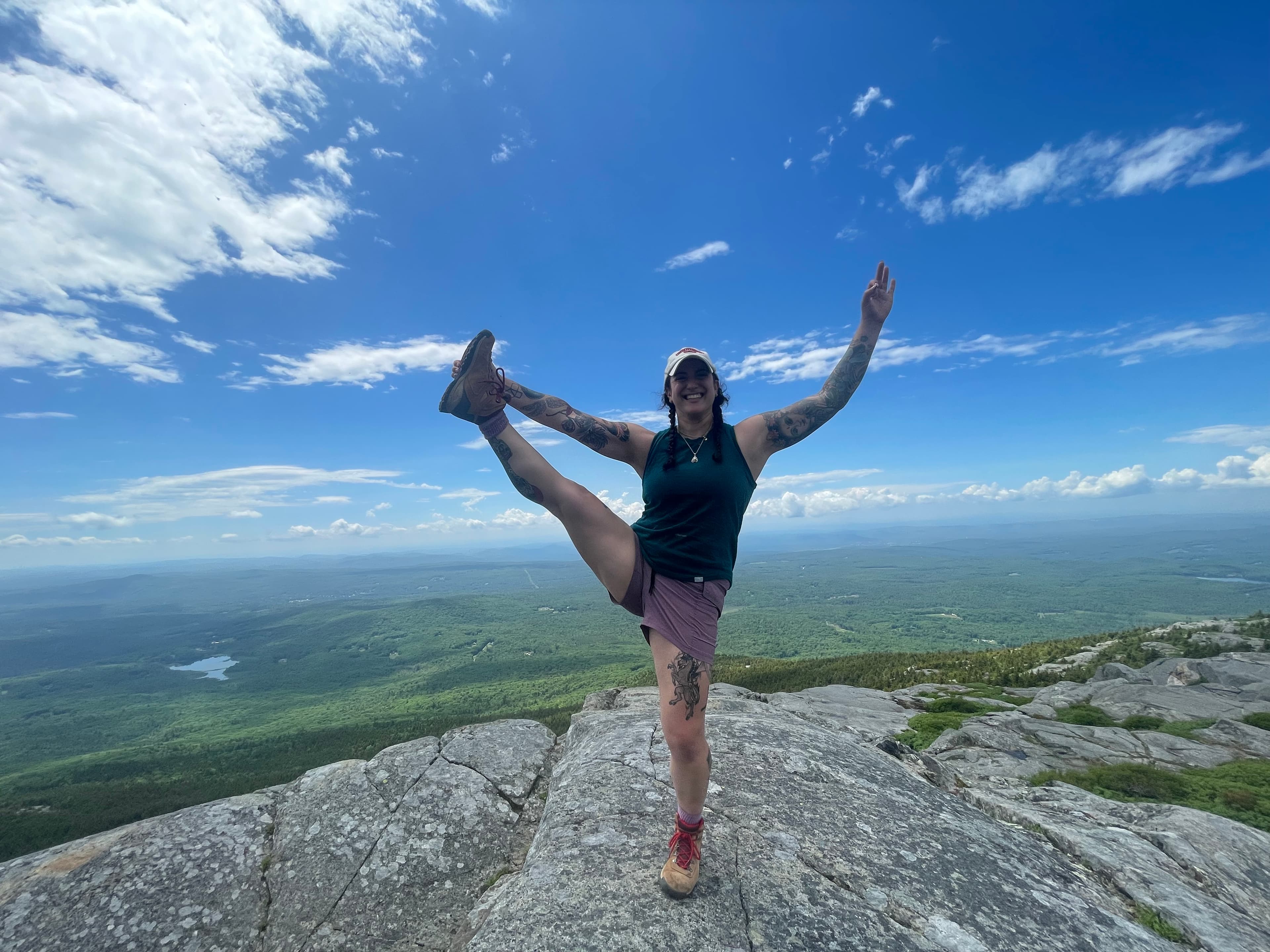 Photo of Jennie smiling doing a yoga pose (Utthita Hasta Padangusthasana or “standing one hand to toe pose”) on top of a mountain.