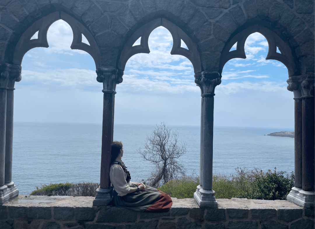 Picture of Tony sitting on a stone wall. They are leaning against a pillar supporting a series of arches, and their head is turned towards the dark sea.