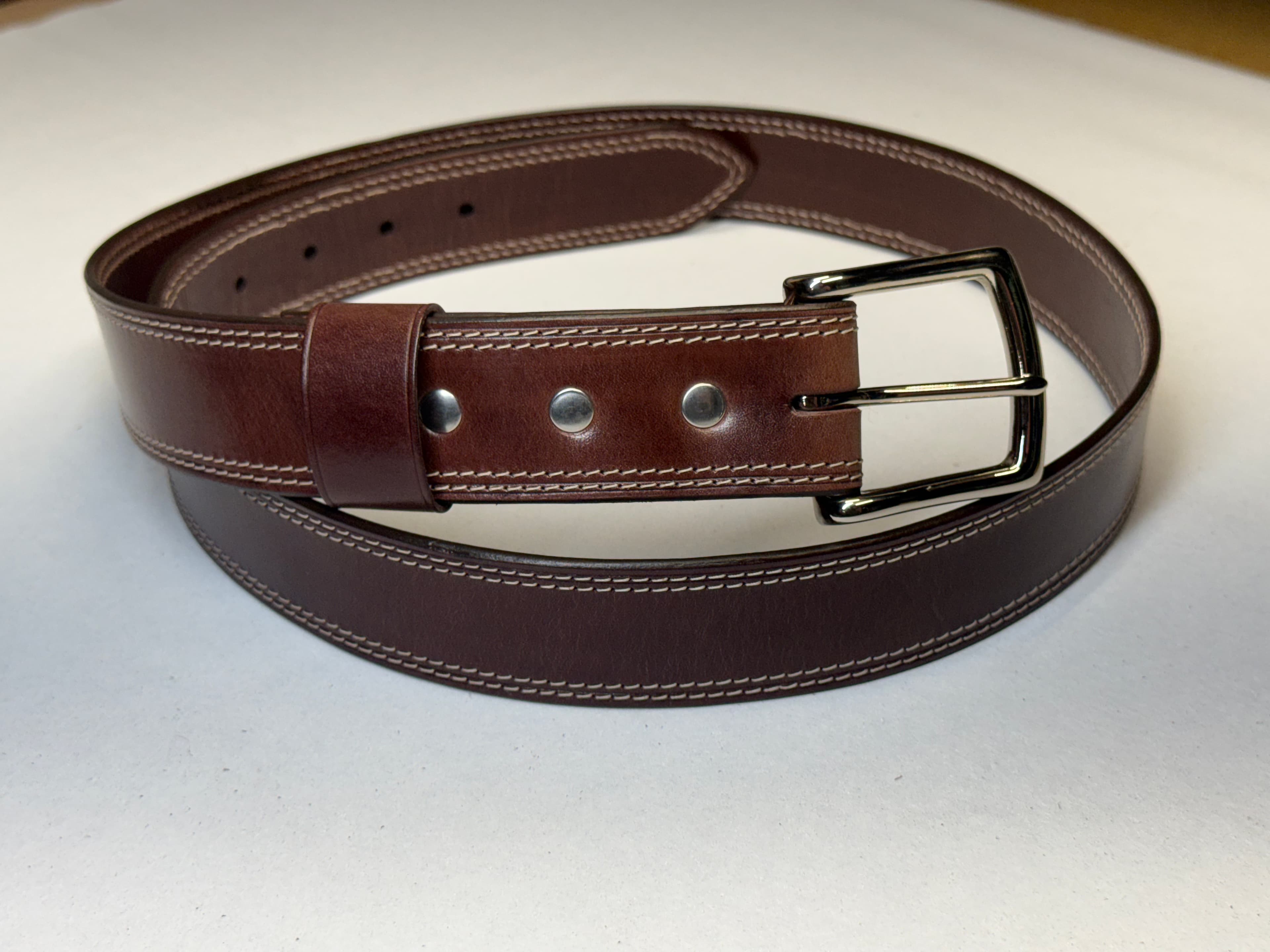 A completed reddish chocolate-brown Maya belt with two rows of off-white stitching along each edge, a silver-colored buckle, and three fasteners.