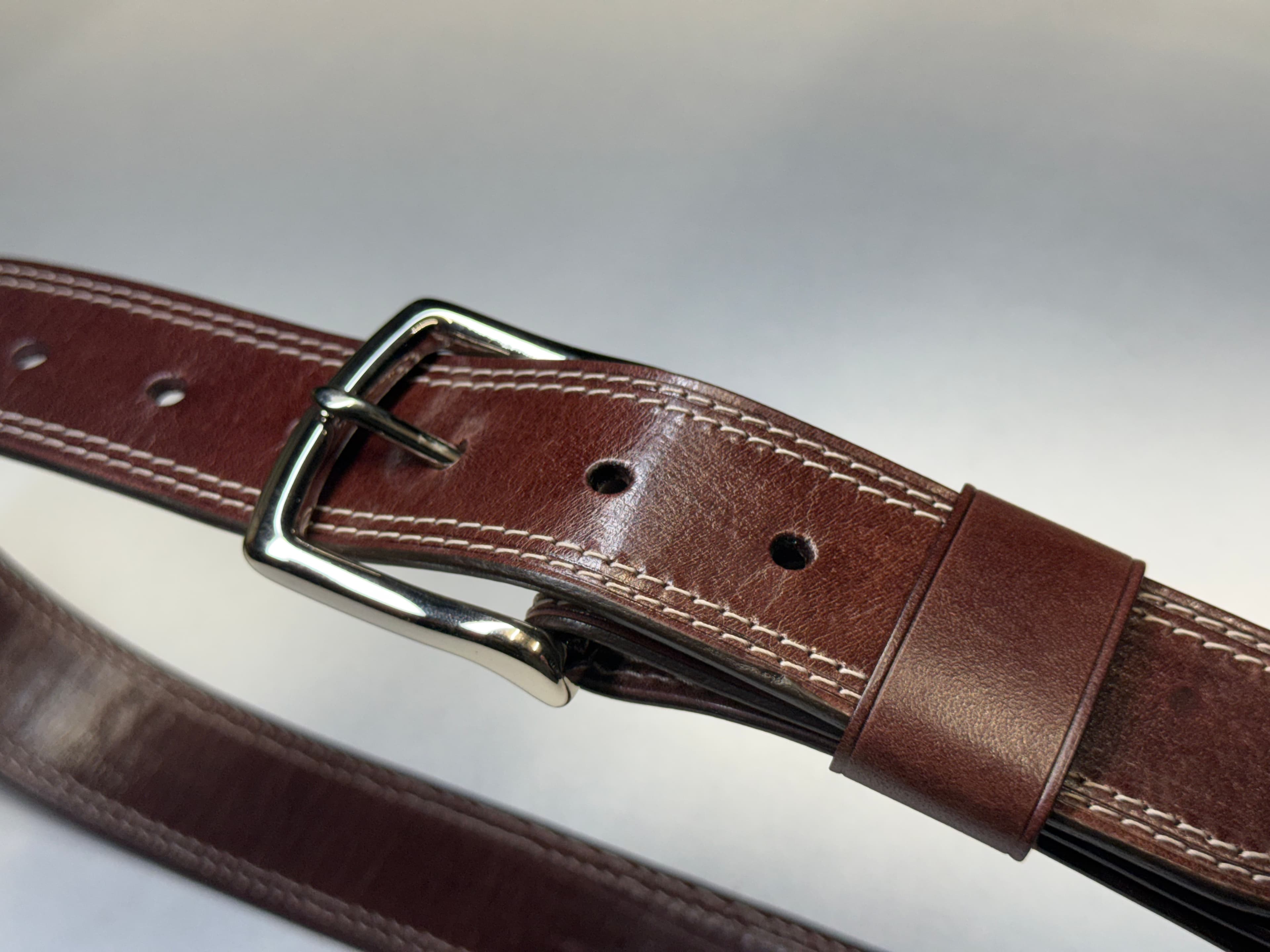 The belt near its buckle and keeper, showing differences in sheen and color, fine pale lines around a sizing hole, and a small irregular area at the bevel.