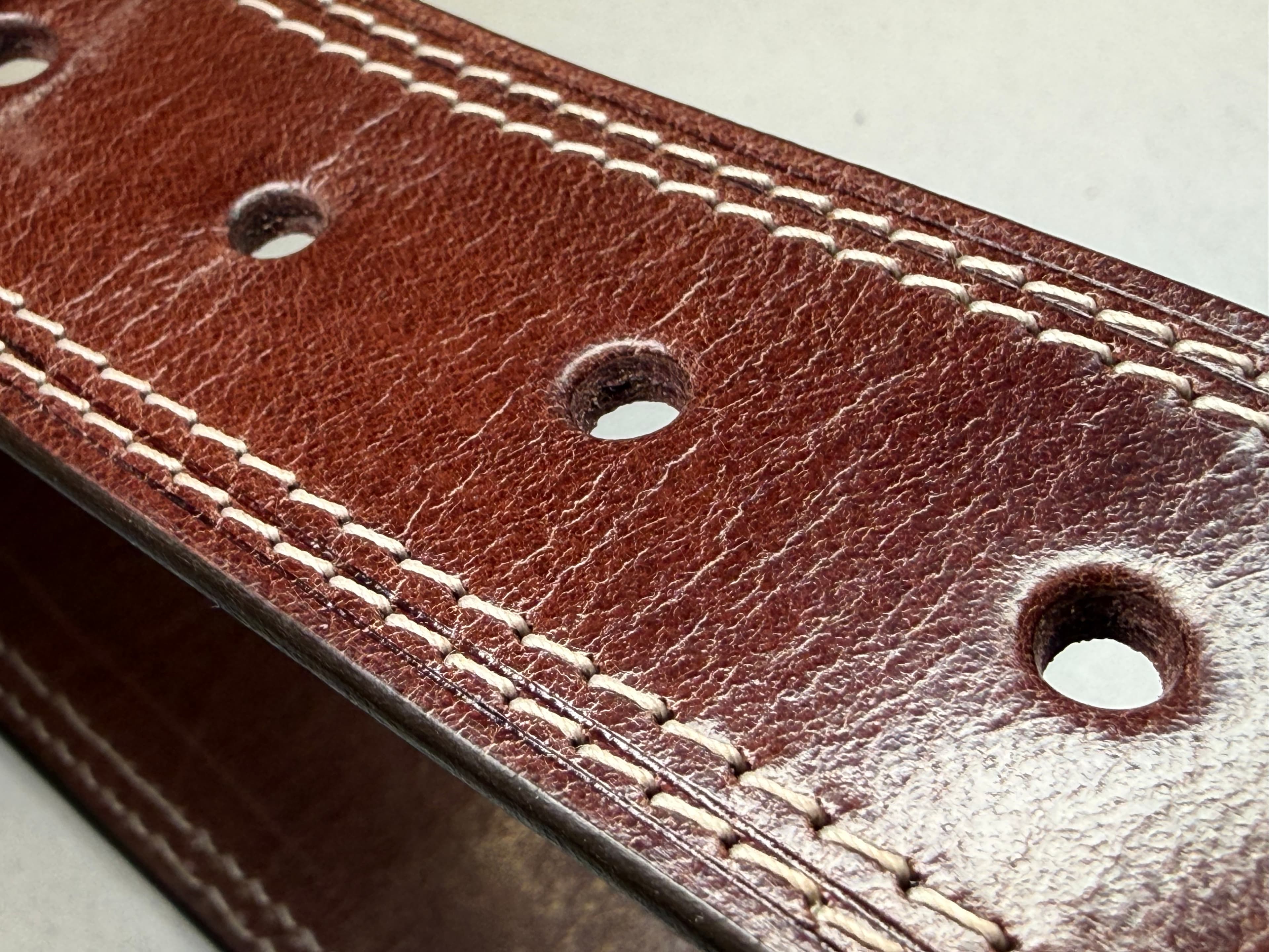 A very close view of the belt’s sizing holes and stitch rows under raking light, revealing a network of fine pale grain lines.