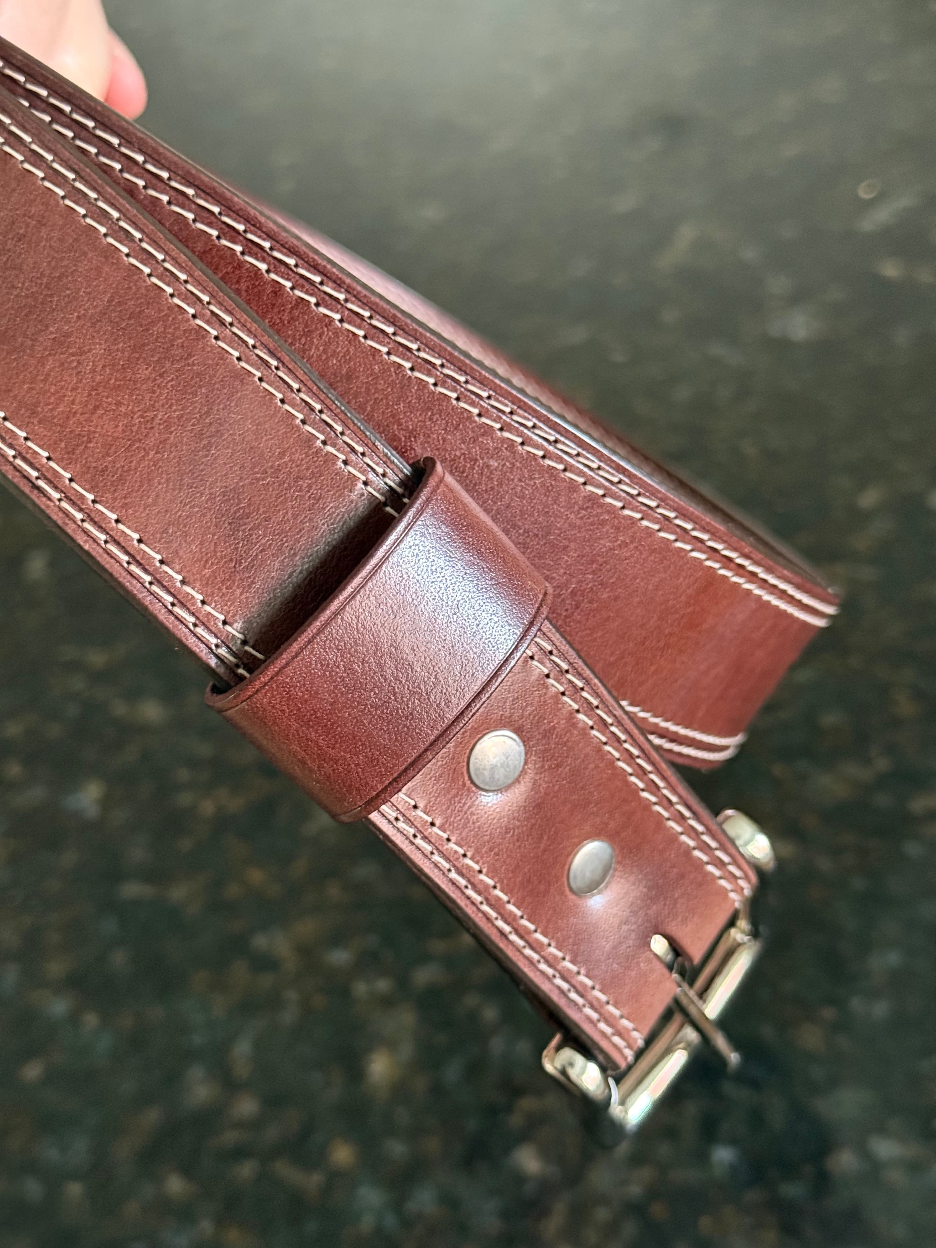 The finished Maya belt held above the granite counter, showing a polished keeper, reddish-brown strap, paired stitch rows, and silver-colored hardware.