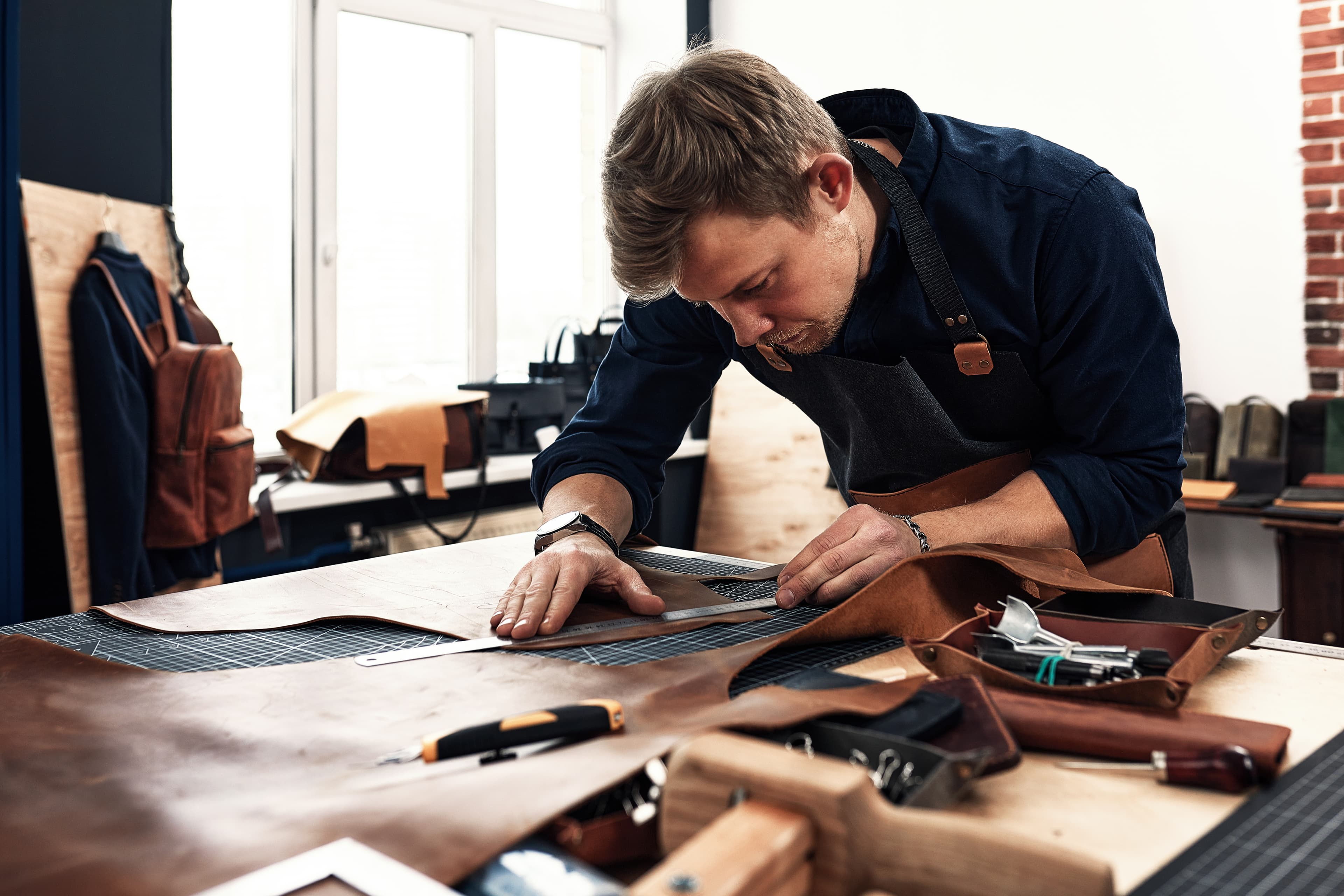 A leatherworker measures and marks patterns on hides at a workshop table.