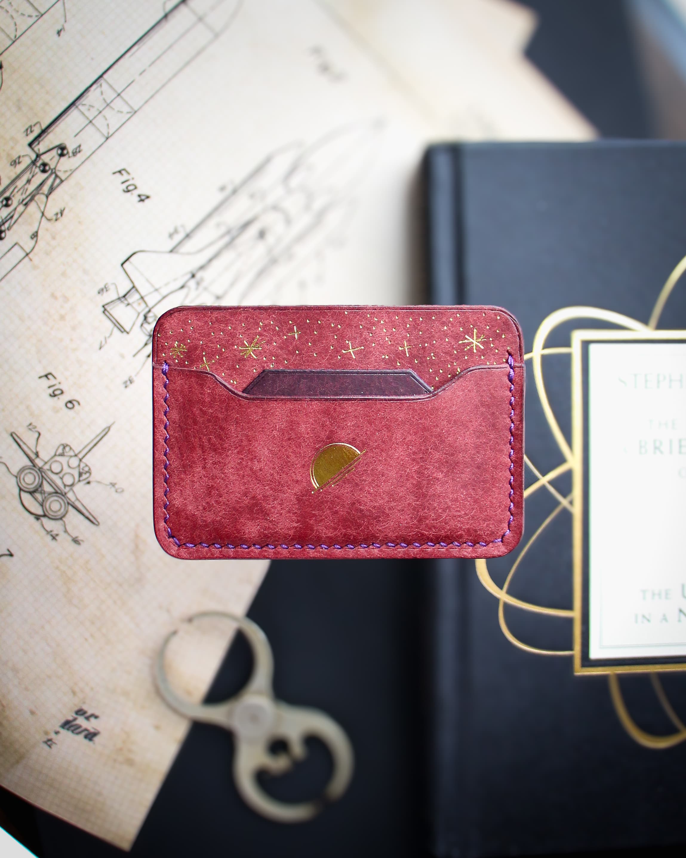 A red leather card wallet displayed with pattern drawings and design books.